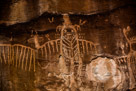 This figure actually has some attributes from other figures previously addressed on this cliff face. Of course there is the owl like 'wings'. But also there is the torso with left and right lines diverging from a central spine. Under these is an area decorated by dots and divided by the continuation of the spine. The bottom of the spine appears to split into a sort of 'tail'. Stubby legs and feet of two 'toes' each are indicated. The head appears to have a 'beard' and wears a head dress with downward directed 'feathers', two on each side.  Eyes and mouth are indicated but no neck. The left and right wings are not connected with a line through the body. There appear to be some birds or insects about the head and wings of this figure. Six dots are on each side of the face and two dots under the right 'wing', three under the left.