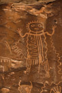 This figure is a good example of what I refer to as the disciplined symmetry and targeted asymmetry of this rock art style. Note the vertical line running from just under the 'mouth' all the way down to separating the legs of this character. About this line the head, arms, hands, legs, feet, head treatment, eyes, neck and torso sides appear purposefully symmetric to the extent possible with the technology. Then inside the torso and around it there are targeted asymmetries. To the right, this section of the torso is divided into three vertical areas by two bold vertical lines with pecking filling the  bottom third of the first vertical column but no further development. To the left there is an elaborate wavy line that fills the left torso cavity, exits the torso and continues the wavy action until it terminates on the figures left elbow. To the figures right there is another wavy line originating on the right elbow and moving down the cliff face to terminate on the right elbow of another figure.