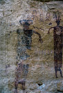 Spalling has damaged the left figure, but it is apparent that the figure originally had a left arm rather similar to the right. There was something to the left of the head. Above the head are six circle figures all with streamers. There appears to be two colors of paint, a deep violet and a near black violet,  used to create the main figure. The eye 'pupils' and mouth/neck are of the deep violet. The main body appears to be made by parallel vertical stripes that totally fill the torso of the figure. The snake is at head/neck level and flicking its tongue at the figure. While the circles at the top(with the streamers) appear similar, the very top figures are more like inverted horseshoes. Actually a pair of inverted horseshoes. Particularly the right two.  The one to the upper right of the main figures head appears to be a combination of the circle with streamers and the horseshoe figures.  When at the site, there appears to be traces of white in the centers of the circles with the streamers.