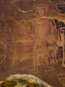 Well this is certainly one of the three Kings. 
But it should be noted that this character is drawn over older petroglyphs. There are several spirils and some sort of sparcely pecked glyph around the thighs. 
This kind appears to have been painted. Probably red is the only remaining color but originally other colors were present.  The King appears to have been totall highlighted in red. Torso, arms, legs, shield, spear, scalp, head and interior details. The king is wearing a elaborate brestplate made of two tiers of shells? and a tunic? covering his upper torso. Rows of beads? define the edge of this tunic? and there is a line especially pecked down the center?.  The kind wears what appears to be a crown that comes down to the hair line but exposes the face. Round pentants are indicated on each ear. Two eyes may be indicated but otherwise the face is currently blank. Originally painted??. The arms of this kind appear to have armbands above the indicated elbows. A beaded belt is indicated. 
In his right hand he is holding a round shield with five 'ribbons' hanging below it. The shield and ribbons appear to be decorated with beads.  The shield is dominated by two broad horizontal stripes, the upper currently white, the lower red. 
Also in the right hand and behind the shield the King appears to hold a spear to which is attached what is deemed to be a scalp.