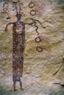 Now this other fellow with the solid feet. He is touching the snake to the right. Is that a bird on the left of his head. If so there are nine such 'bird's over his head. And the 'antennae' on his head are rather like the antennae of moths. The four large hollow circles with streamers are presented with the streamers all to the right. The smaller hollow circles between the figures had streamers to both the left and right.  This snake also has its tongue out pointing to the head of the figure and at about head/neck level.  The single large 'bird' figure is directly opposite.  The main figure appears to be created using vertical stripes and is completely filled except for a circular decoration on the chest. The decoration appears to be made of three concentric circles as opposed to a spiral. Significant??